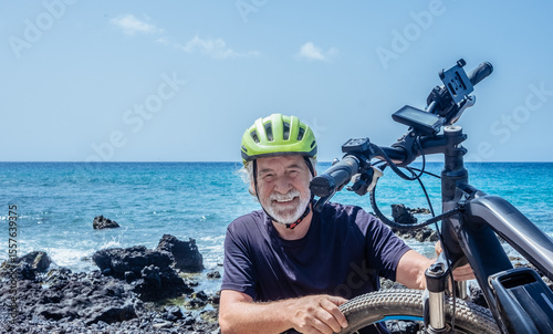 Smiling elderly cyclist man wearing helmet enjoying a sunny day with his bicycle on a rocky beach in a sunny day. Healthy retirement lifestyle. Horizon over water