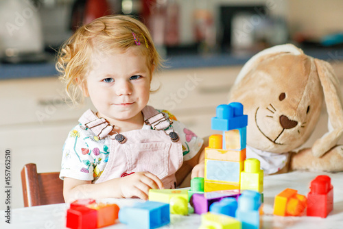 Adorable toddler girl with favorite plush bunny playing with educational toys in nursery. Happy healthy child having fun with colorful different plastic blocks at home. Cute baby learning creating.