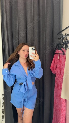 young girl teenager trying on a suit in a fitting room blue shirt buying new things shopping taking pictures of herself in the mirror with her phone purchases spending money
