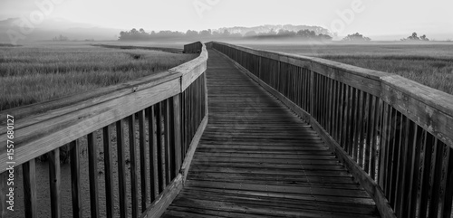 A black and white photo of a boardwalk over a salt marsh with mist hanging over the marsh grass and trees in the distance