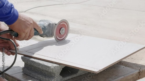 Construction site scene with a worker in blue clothing using an angle grinder to cut a gray tile, highlighting detailed craftsmanship and precision in tile preparation.