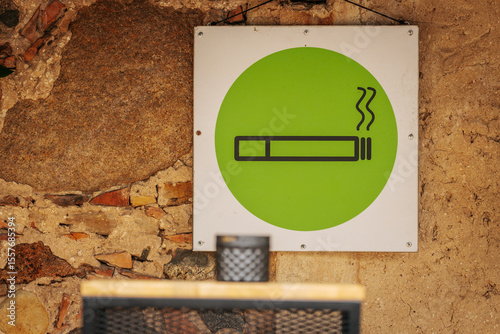 A green smoking area sign with a cigarette icon is mounted on a rustic stone wall, indicating a designated place for smokers.