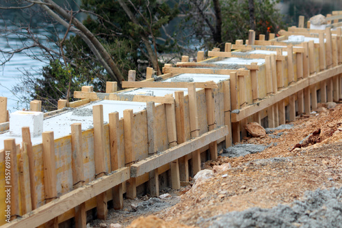 New concrete is setting inside sturdy wooden formwork, creating a straight foundation line for a riverside path, surrounded by soil and sparse vegetation