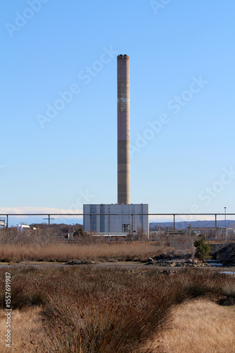 Tall concrete smokestack towers over a factory complex and grassy field, representing industrial presence and air emissions against a clear blue sky