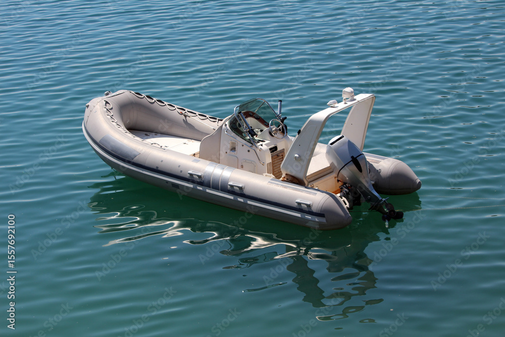Naklejka premium A sleek inflatable motorboat with steering console and outboard engine floats alone on tranquil blue water, reflecting sunlight and gentle waves