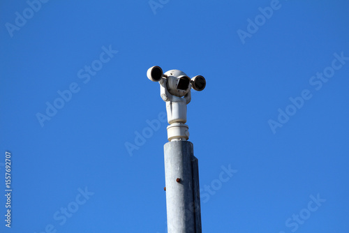 A trio of security cameras mounted on a high metal pole points in different directions, monitoring the area beneath a clear, vibrant blue sky