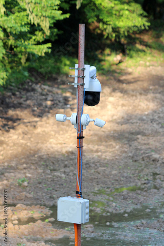Multiple security cameras and electrical box are installed on a rusty pole, monitoring a forest clearing with dense trees in the background