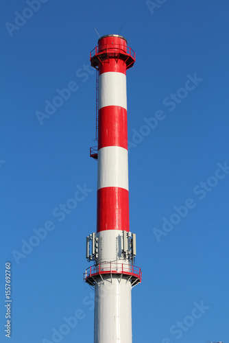 High industrial chimney with bold red and white stripes rises against a clear blue sky, equipped with antennas and safety railings for modern industry