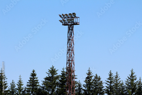 A towering metal floodlight structure rises above dense spruce trees picea abies, set against a cloudless blue sky, blending technology and nature