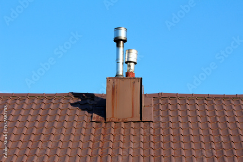Two modern metal chimneys rise from a brown tiled roof, sharply outlined against a cloudless blue sky, showing a blend of utility and architectural style