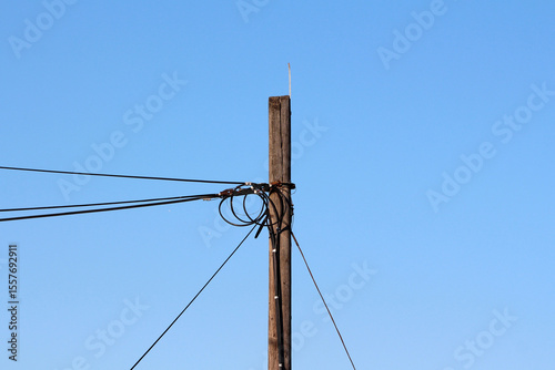 A weathered wooden utility pole supports a web of tangled electrical wires, silhouetted against a bright blue sky, highlighting rural infrastructure and energy distribution