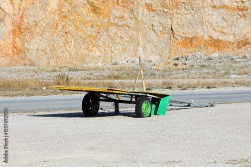 Yellow flatbed trailer with a green wheel and wooden frame sits on gravel beside a steep orange quarry wall, highlighting rugged construction and industrial surroundings in sunlight