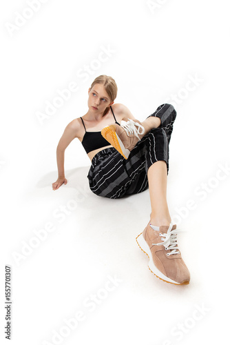 A model sits casually on a white surface, wearing a black top and striped pants while showcasing trendy sneakers. The relaxed pose enhances the outfit's modern vibe in a fashion setting.