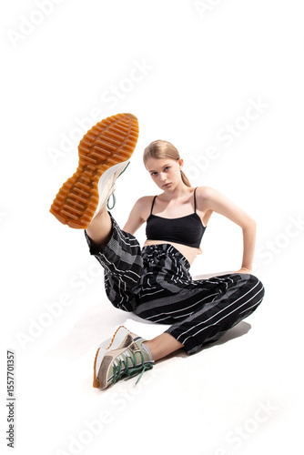 A young model poses with one leg lifted and a confident expression, highlighting her sporty outfit and trendy sneakers during a fashion shoot against a plain background.