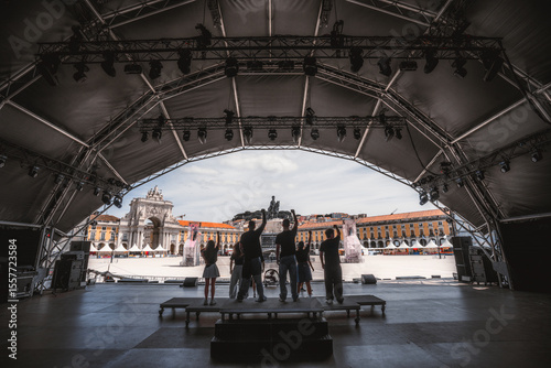 Wide-angle view from behind of a group of people standing on an outdoor stage during Pride event, raising fists toward the iconic Arco da Rua Augusta in unity and celebration