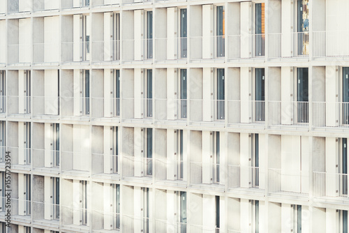 Side view of a modern apartment or hotel building with minimalist white balconies, glass doors, and repeating geometric architecture, illuminated by soft natural daylight and casting subtle shadows