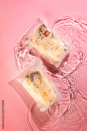 wo sealed packets of colorful food ingredients float gently in a rippling water pond on a soft pink background, creating a vibrant contrast. The ingredients within the packaging seem to be a blend of 