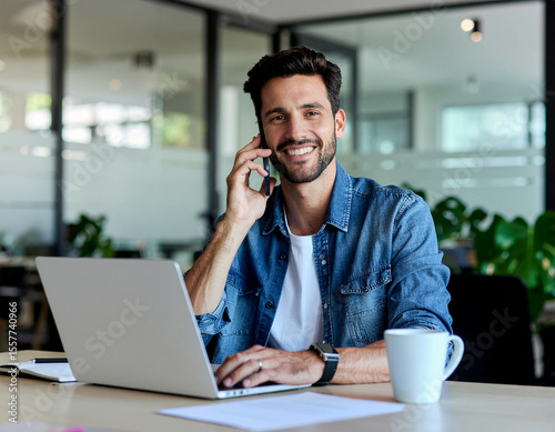 Portrait of young smiling cheerful entrepreneur in casual office making phone call while working with laptop