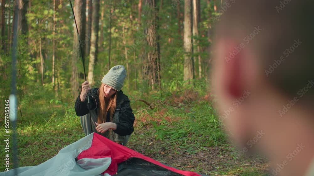 Blurred head appears in foreground observing young girl squatting and fixing tent fabric hook on flexible pole during camping setup in forest