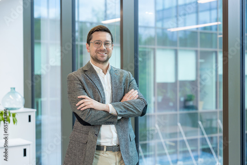 A smiling young man, founder and owner of a firm and company, stands in the lobby of an office building, arms crossed on his chest and confidently looking into the camera