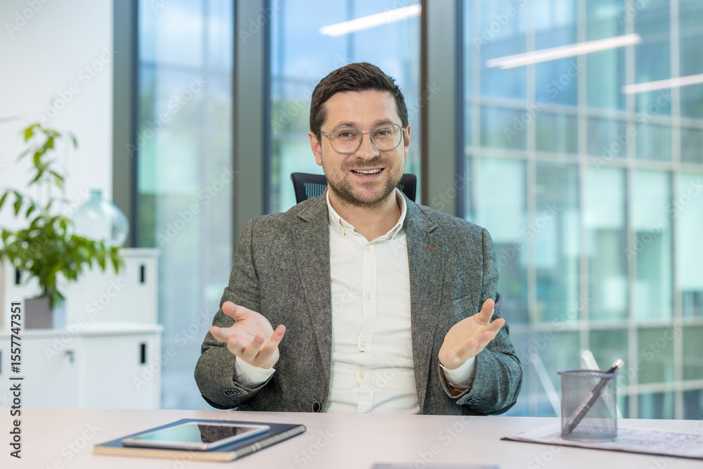 Fototapeta premium Smiling young male businessman sitting in the office at the table and holding an online conference via video call, gesturing with his hands at the camera