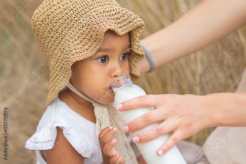 Obraz na płótnie A large portrait of a little mulatto girl who drinks milk from a bottle held by her mother's hands at a picnic in the field