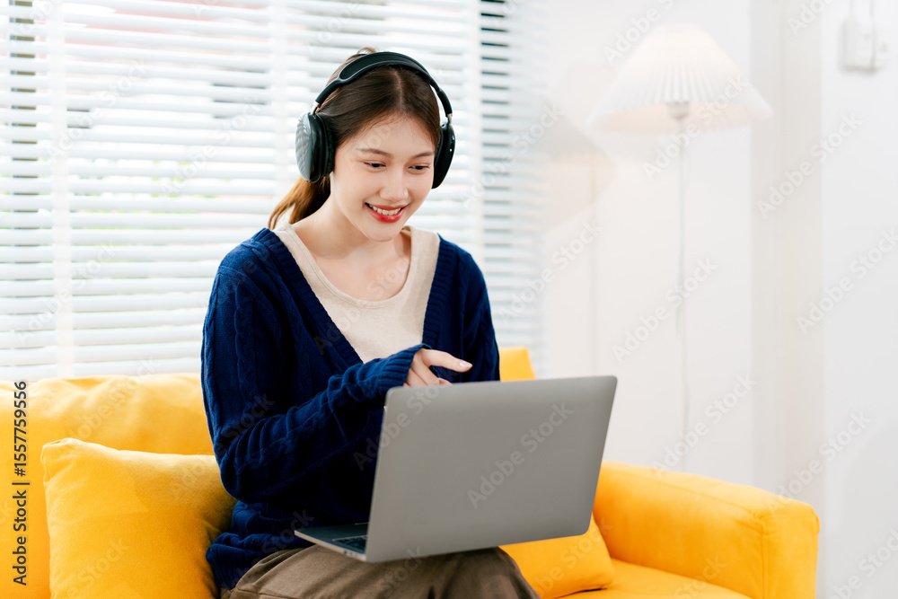 Naklejka premium Beautiful Asian female student studying online at home. Young woman wearing earphones listening to music online.