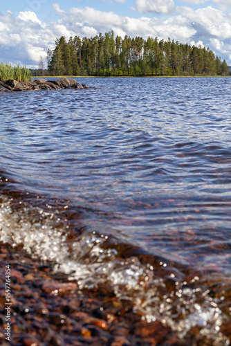 Siljan lake clear water pebbles shore waves, in Isunda, Dalarna, Sweden. Isundaholmen island with forest trees. Authentic spring Nordic environment sunshine daylight background vertical scene.