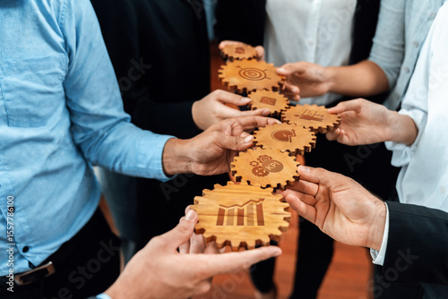 A diverse business team collaborates by holding wooden gears, each featuring icons that represent various performance metrics and strategic goals. Amity