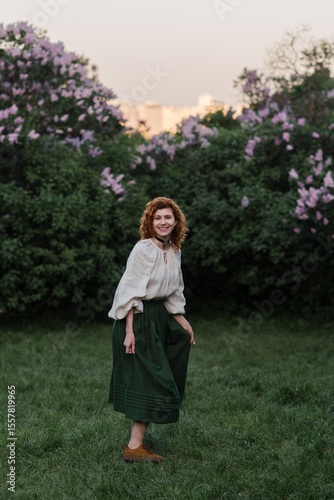 Young pretty caucasian woman in romantic outfit walking in blooming spring garden. Artistic outdoor photo of beautiful red-haired girl posing in fragrant lilac blossom.