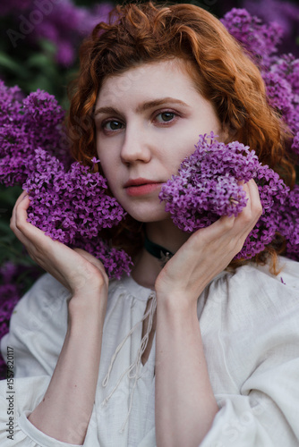 Young pretty caucasian woman in romantic outfit walking in blooming spring garden. Artistic outdoor photo of beautiful red-haired girl posing in fragrant lilac blossom.
