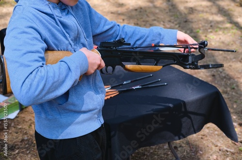 Canvas Print Teenager in blue hoodie loading black sport crossbow at shooting range in forest
