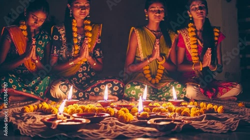 Indian women in traditional saris praying together with joined hands near lit diyas and yellow flower garlands during diwali festival celebration