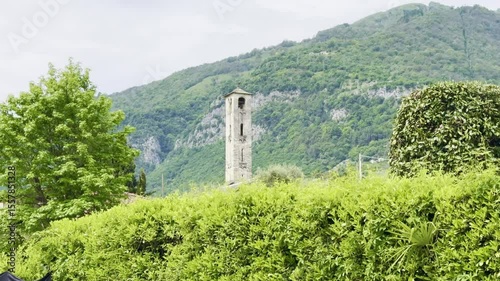 Ancient stone bell tower of Chiesa di Sant’Andrea standing tall among lush greenery with dramatic mountain backdrop, medieval Romanesque architecture near Lenno, Lake Como, Lombardy, northern Italy