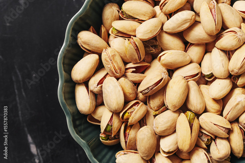 Pistachio nuts in green bowl. Healthy roasted pistachios with shells on black marble background. Crunchy vegan snack or ingredient. Natural food closeup from above. High protein vegetarian option.