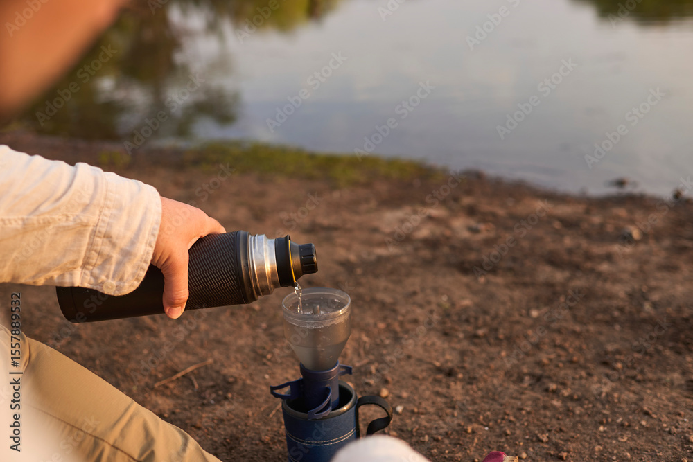 Fototapeta premium Coffee filtering process. Hispanic man pouring hot water, making coffee for breakfast outdoors early in the morning by a creek, in El Palmar National Park, Entre Rios, Argentina.