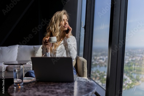 A young and ambitious woman enjoys an engaging phone call while savoring a rich cup of coffee and diligently working on her laptop by a large window that offers a stunning view of the city skyline