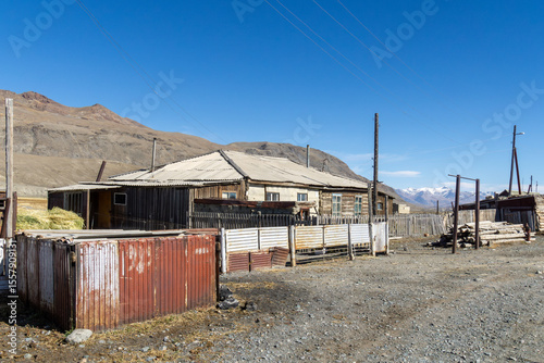 Rustic wooden house surrounded by fences and outbuildings in a remote mountainous area, showcasing rural architecture and natural landscape under a clear blue sky