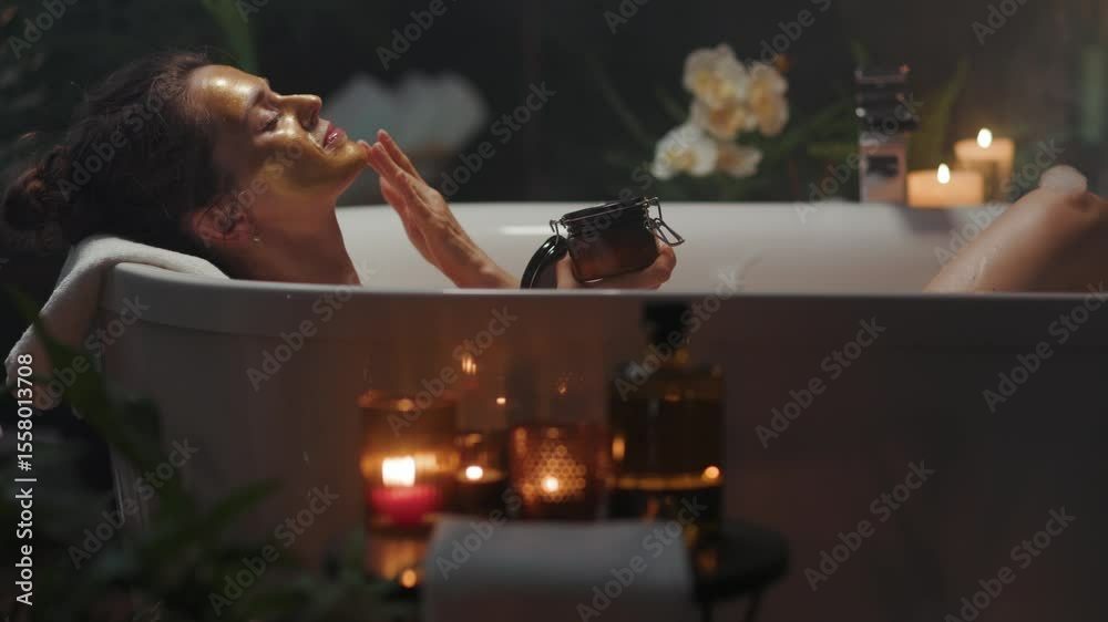 A serene woman in her 30s, adorned with a gold face mask, gently touches her cheek while relaxing in a candlelit bathtub, surrounded by lush plants, creating a luxurious beauty and wellness experience
