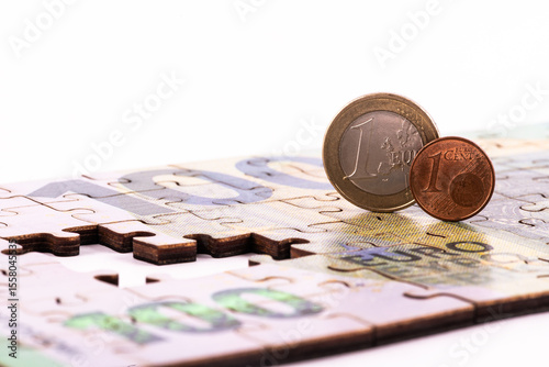 A 100 euro note made from puzzle pieces, 1 euro and 1 euro cent coins on a white background