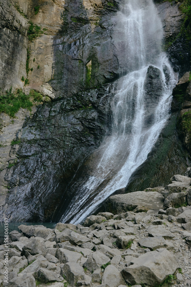 Fototapeta premium Natural waterfall cascading down rocky cliff surrounded by green moss and rugged stones under diffuse daylight in a serene mountainous landscape.