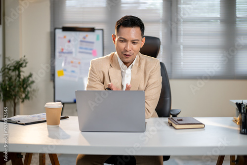 Asian businessman excitedly talking or explaining on video chat via laptop at office's working table.