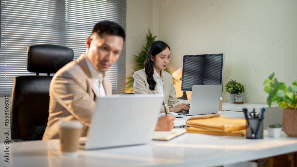 Fototapeta premium Businessman writing with pen in notebook and looking at laptop on office table aside female coworker