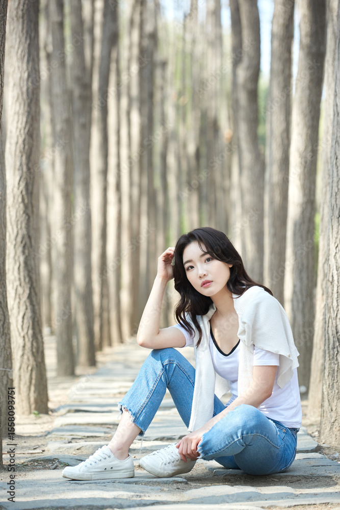 Young Female Sitting Relaxed in Tree-Lined Park Pathway Wearing Casual Outfit Young Female Sitting Relaxed in Tree-Lined Park Pathway Wearing Casual Outfit