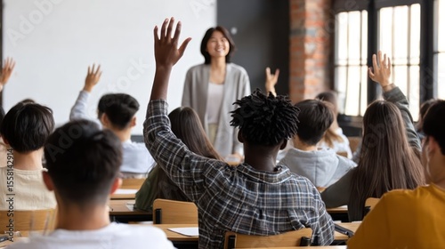 Diverse group of Gen Z Asian high school students in modern classroom, one Black male student raising hand to ask question to senior female teacher during interactive lecture