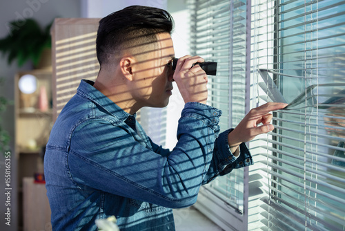 excited man spying through the window blinds with binoculars