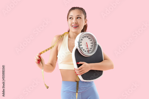 Sporty young Asian woman with tape measure and weight scales on pink backgrou...