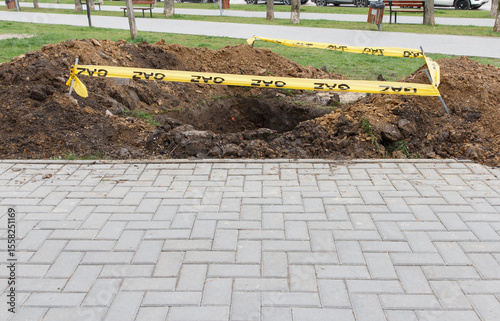 Deep hole surrounded by dirt and yellow caution tape on grassy area near paved sidewalk