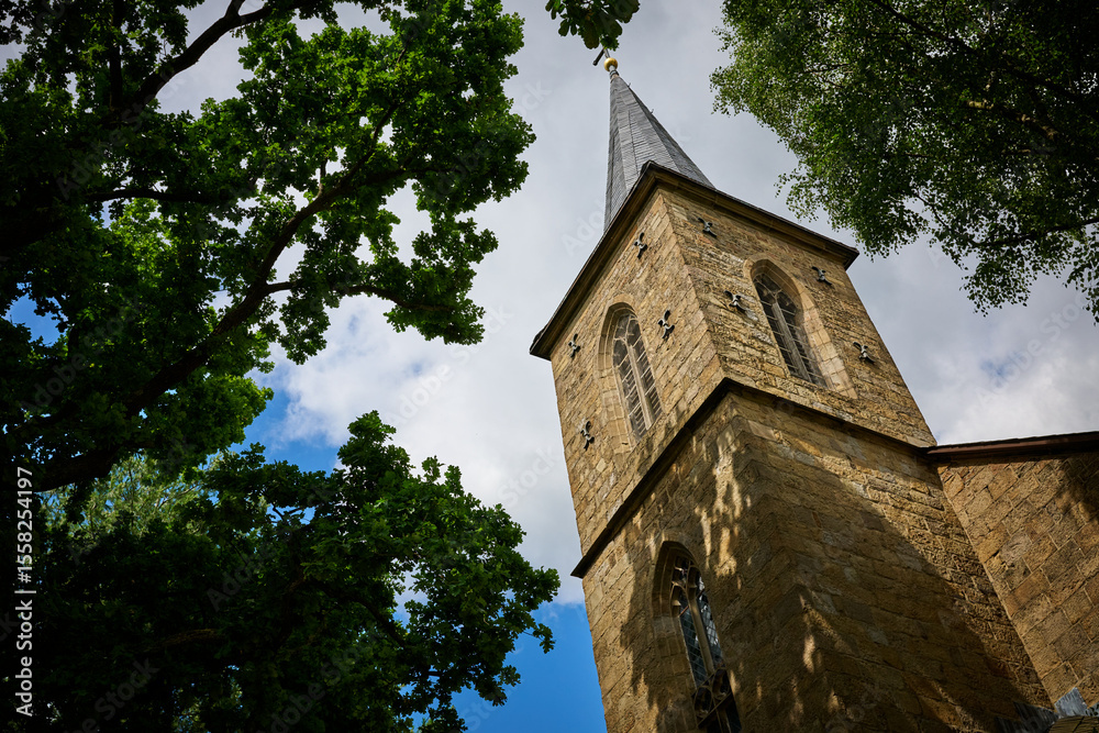 Historic Stone Church Tower in the German city of Hattingen Framed by Lush Green Trees Against a Partly Cloudy Blue Sky on a Bright Historic Stone Church Tower in the German city of Hattingen Framed by Lush Green Trees Against a Partly Cloudy Blue Sky on a Bright