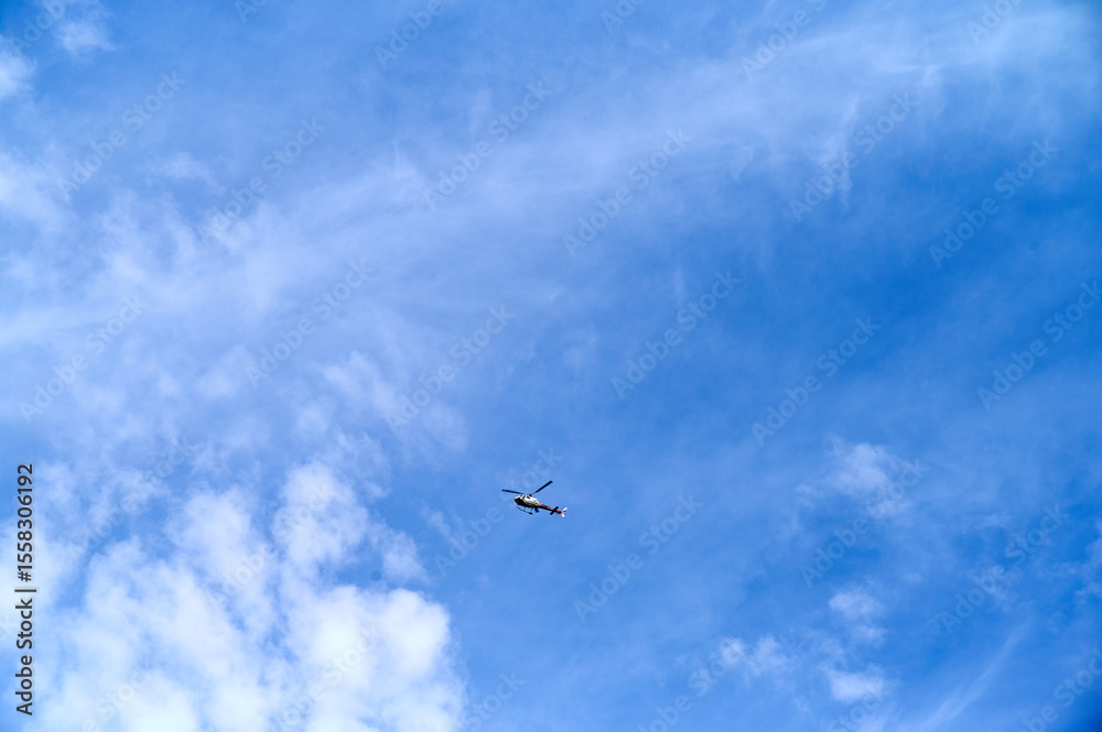 Fototapeta premium A helicopter soars through a clear blue sky, surrounded by light, fluffy clouds, showcasing a vibrant day.